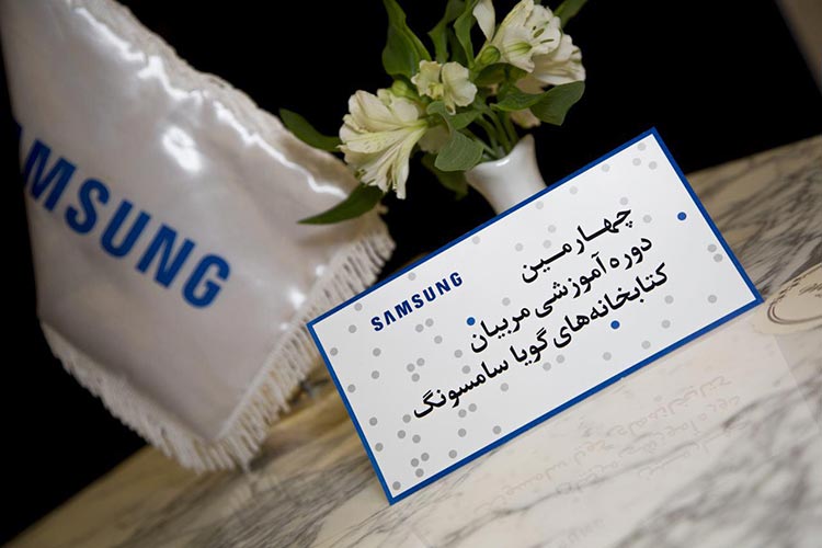Printed Samsung Audio Library tabletop sign with Persian event typography and grey dots inside a blue frame.